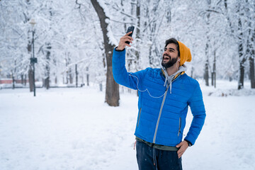 Excited young man using mobile phone, having video call or taking selfie while standing in public park on snowy cold winter day.