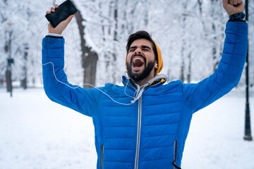Young cheerful man raising arms up and singing with earphones and smartphone in his hand. Young handsome man in blue jacket expressing excitement and joy while listening music over cable earphones.