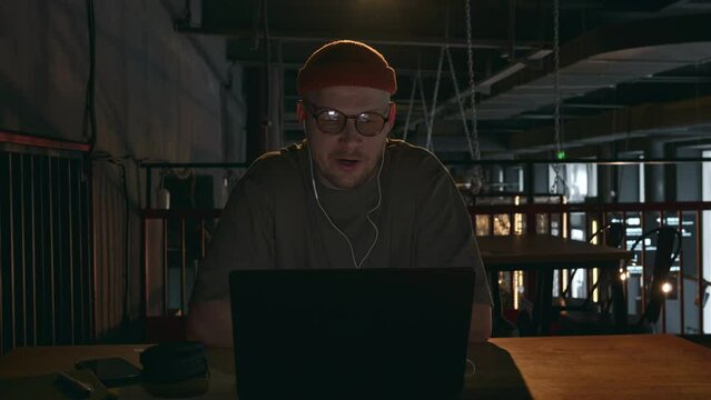 A male Internet scammer communicates via video link. He waves amiably while sitting in front of a laptop in a dark room with headphones on. High quality 4k footage