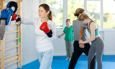 Fototapeta na wymiar Focused woman in boxing gloves practices punches with man in mitts in gym