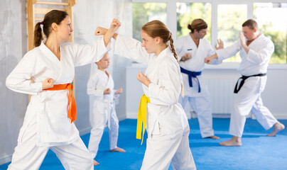 Fototapeta na wymiar Focused teen girl and woman in kimono practices combat technique of punching and blocking at family martial arts class