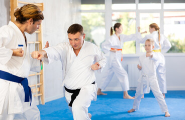 Focused man in kimono practices combat technique of punching and blocking with trainer at family martial arts class