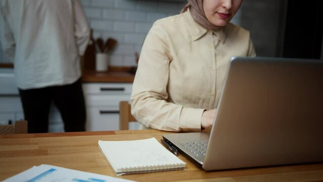 Close up happy young couple at home in kitchen with freelancer woman working on laptop, writing on notebook paper at kitchen counter being supported by her husband