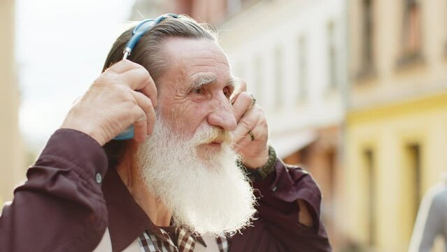 Happy Bearded Senior Old Man Wearing Wireless Headphones Choosing, Listening Favorite Energetic Disco Rock N Roll Music Dancing Outdoors. Portrait Of Elderly Grandfather Walking In Urban City Street