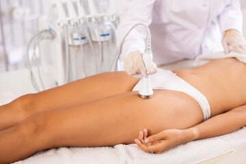 Fototapeta premium Aesthetician administering hardware procedure on female patient in cosmetology clinic, using high frequency ultrasound attachment to reduce body fat and contour thighs