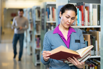 Portrait of a focused asian woman standing in a public library near the bookshelves and reading selected literature