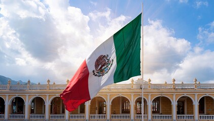 Plano general de bandera de México ondeando en lo alto con un cielo nublado y destello de luz detrás - obrazy, fototapety, plakaty