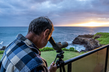 Mature Caucasian man by the ocean using a spotting scope to watch whales and dolphins as the sun...