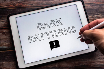 Keyword “Dark Patterns” seen on tablet on wooden table. Man hand holding wireless stylus pen. Deceptive online business practice concept - obrazy, fototapety, plakaty