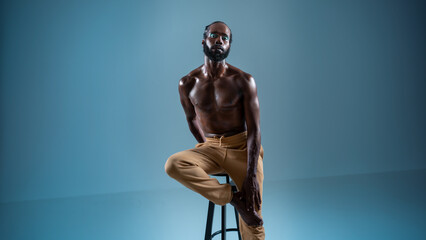 Fototapeta na wymiar Black bearded gay wearing make-up sitting chair isolated on blue. African-american gay muscular shirtless man wearing make-up looking at camera. 