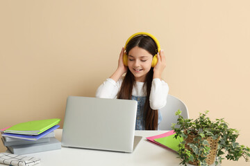 Little girl in headphones with laptop doing lessons at table on beige background