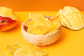 Wooden bowl with slices of dried mango and fresh fruit on orange background