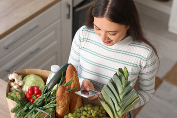 Young Asian woman with mobile phone and shopping bags full of fresh food at table in kitchen