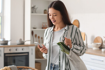 Young Asian woman with eco bag full of fresh food and mobile phone in kitchen