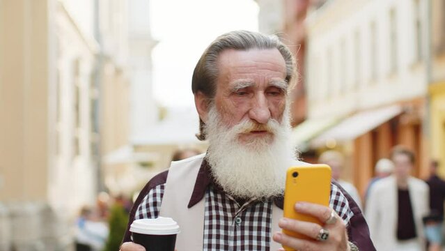 Happy senior old man use smartphone typing text messages browsing internet social media web app working chatting online enjoying morning to-go coffee hot drink. Grandfather tourist walking in street