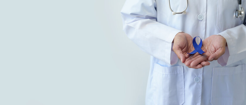 An image of a woman's hand in a doctor's uniform and stethoscope holding a blue ribbon represents fighting for life. Treatment from colon cancer World cancer day concept, copy space, banner, panorama