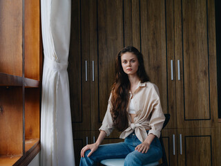 Woman sitting on a chair in home interior and simple clothes near the window, comfort and coziness, lifestyle relaxation on the weekend, pensive look of a beautiful girl.