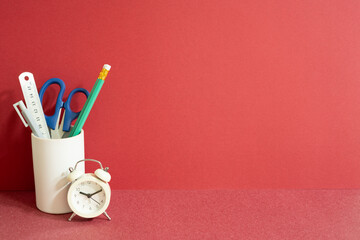 Pencil holder with ruler, scissors, pen and alarm clock on red desk. red wall background, workspace