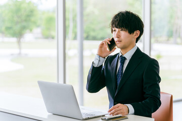 スマートフォンで話しながらパソコンを使う若いアジア人ビジネスマン
