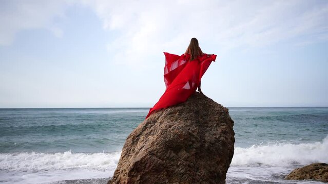 Red dress sea woman. Happy woman with flowing hair in a long flowing red dress stands on a rock near the sea. Travel concept, photo session at sea