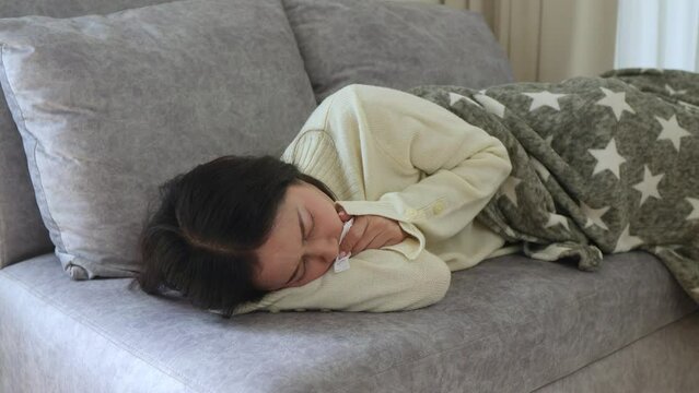 Attractive young woman wrapped in warm blanket, sits on comfortable sofa at home, blowing her nose in disposable tissue. Allergy to dust, animal dander, cold, or viral infection