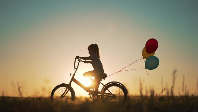 Dream kid. Silhouette of kid on bike in park. Girl rides in park on green grass. Child games in nature.Traveling with balloons on bike.Active child freedom in summer.Girl learns to ride bike in nature