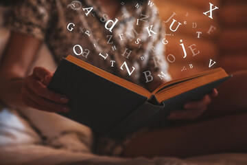 Woman reading book with letters flying over it on bed at home, closeup