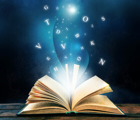 Open book with magic light and glowing letters flying out of it on wooden table against black...