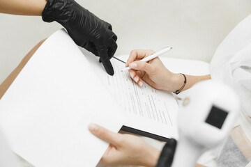 female client signs consent form while sitting during medical consultation with cosmetologist....