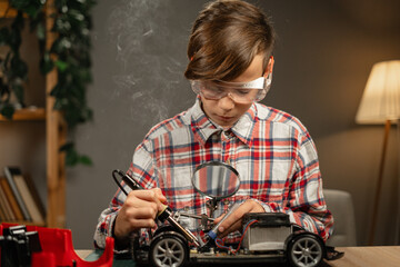 A child in safety glasses soldering wires and chips in a toy car while sitting at home in the...