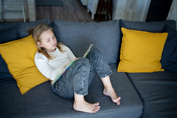 A pretty junior schoolgirl enjoys reading a book, completely immersed in its contents, on the couch...