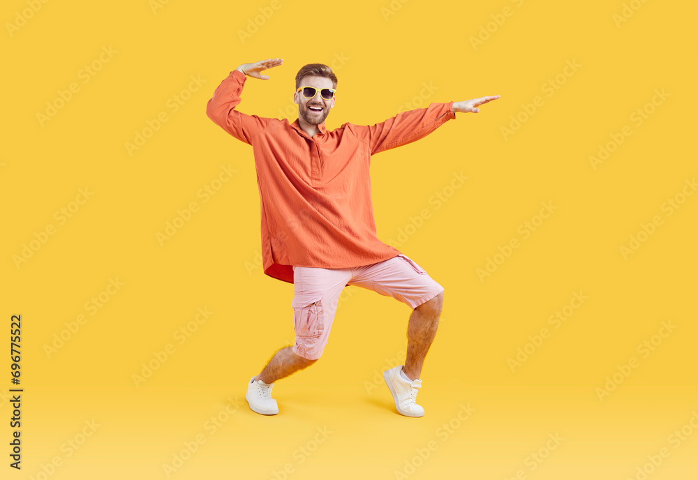 Wall mural Happy guy in casual summer clothes having fun in fashion studio. Full body length shot of funny young man in orange shirt, pink shorts, white sneakers and sunglasses dancing on yellow color background - Wall murals