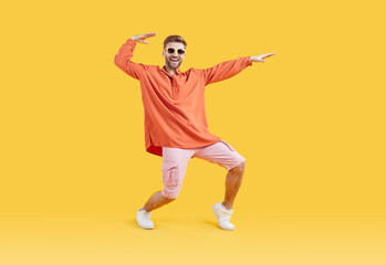 Fototapeta na wymiar Happy guy in casual summer clothes having fun in fashion studio. Full body length shot of funny young man in orange shirt, pink shorts, white sneakers and sunglasses dancing on yellow color background