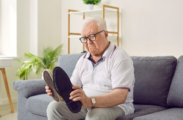 Senior man with dementia holding different shoes in his hands. Old elderly person suffering from Alzheimer's disease sitting on sofa at home alone sad and upset. Mental disorders concept. - obrazy, fototapety, plakaty