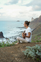 Young long hair man in casual attire, plays a ukulele on a cliff edge, overlooking a tranquil ocean and rugged cliffs
