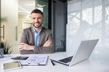 Portrait of successful financier accountant at workplace inside office, senior experienced businessman smiling looking at camera with crossed arms, working with papers sitting at table with laptop. - obrazy, fototapety, plakaty