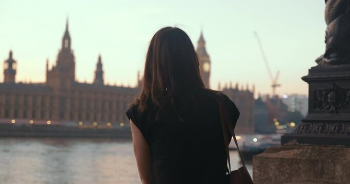Happy attractive young female enjoys view of Big Ben and the Thames river during beautiful sunset. View from the back.