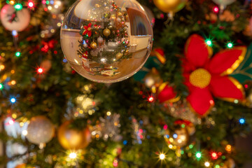 Close-up of reflection of Christmas tree in a silver glass ball, decoration with golden baubles, small colored lights, poinsettia on blurred background, Christmas concept at home. Espace for text