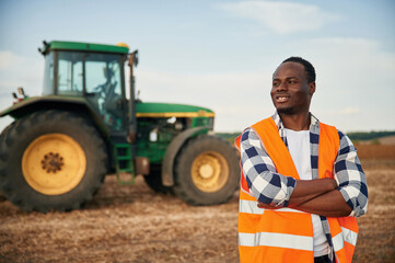 Near the tractor in orange uniform. Beautiful African American man is in the agricultural field