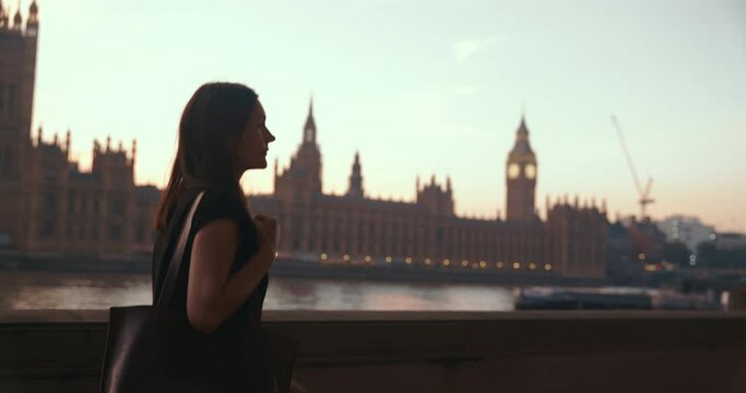 Camera follows young attractive brunette walking along the Thames river in London. Big Ben in the background.