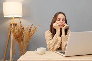 Browsing manager business. Sad bored brown haired woman wearing beige sweater looking on computer display with boring displeased expression while sitting at table in home office