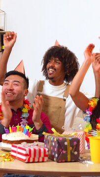 Multi-ethnic group of friends at a birthday party on the sofa at home with a cake and gifts, smiling placing the happy brithday candles