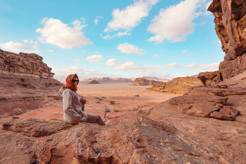 Young woman sitting on rocky ground in desert landscape looking over her shoulder, small vehicle far distance. View from behind. Wadi Rum, Jordan - obrazy, fototapety, plakaty