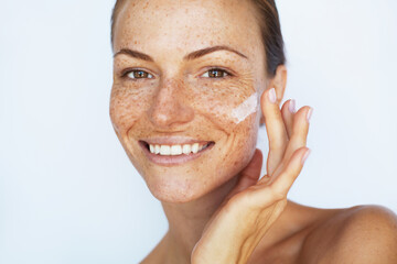 Beautiful woman with a freckles is applying a facial skincare cream and smiling. Beauty skin care, ...