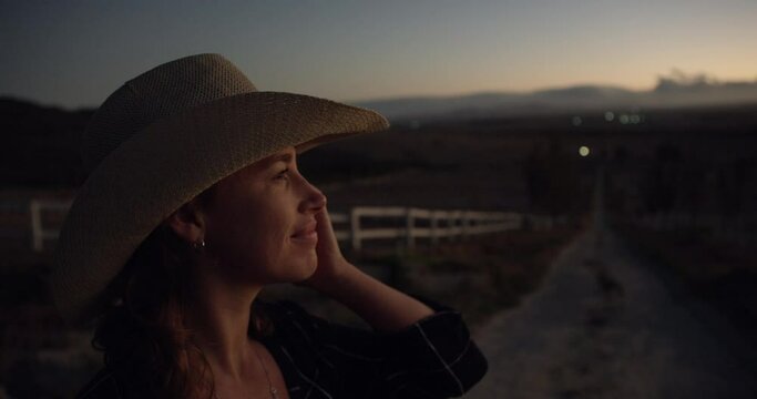 Happy woman, cowgirl and hat at night on farm for sunset, adventure or farming in the countryside. Face of female person or farmer smile for late evening view, natural scenery or outdoor nature