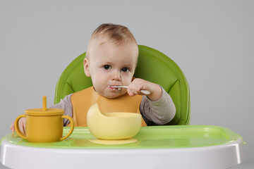 Cute little baby eating healthy food in high chair on gray background