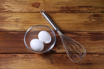 Metal whisk and eggs in bowl on wooden table, flat lay