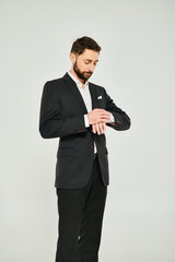 Obraz na płótnie Canvas good-looking bearded businessman adjusting sleeve of black suit on grey backdrop in studio