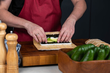 Close up of sushi chef hands making Japanese food. Man cooking sushi at restaurant or at home. Traditional asian rolls on cutting board. Maki sushi with cheese and vegetables: pepper and avocado