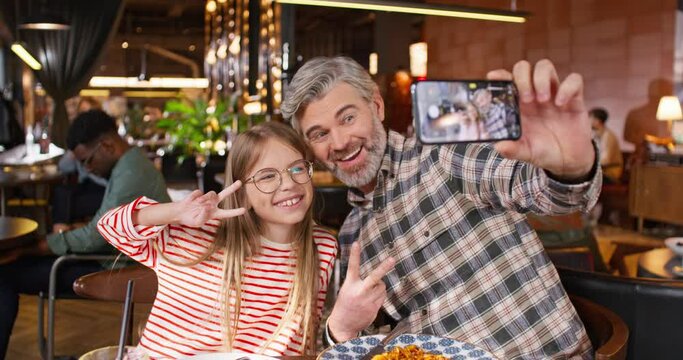 Zooming out from Caucasian happy family resting together in cafe or pizzeria. Handsome father making selfie with his daughter while holding his smartphone. In background clients sitting at their table